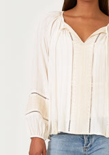 [Color: Cream/Natural] A close up front facing image of a brunette model wearing a cream bohemian fall blouse with embroidered details and sparkly metallic thread. With long raglan sleeves, a split v neckline with tassel ties, lattice trim, and a relaxed fit.