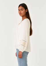 [Color: Cream/Natural] A side facing image of a brunette model wearing a cream bohemian fall blouse with embroidered details and sparkly metallic thread. With long raglan sleeves, a split v neckline with tassel ties, lattice trim, and a relaxed fit.