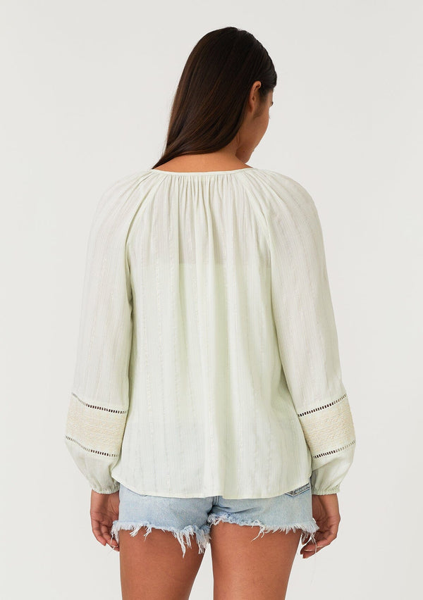 [Color: Mint/Natural] A back facing image of a brunette model wearing a mint green bohemian fall blouse with embroidered details and sparkly metallic thread. With long raglan sleeves, a split v neckline with tassel ties, lattice trim, and a relaxed fit.