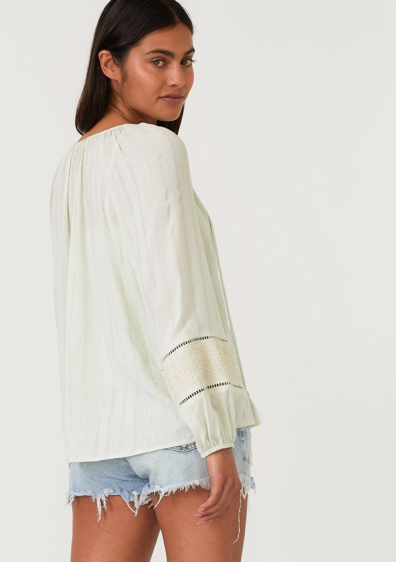 [Color: Mint/Natural] A half body back facing image of a brunette model wearing a mint green bohemian fall blouse with embroidered details and sparkly metallic thread. With long raglan sleeves, a split v neckline with tassel ties, lattice trim, and a relaxed fit.