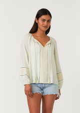 [Color: Mint/Natural] A front facing image of a brunette model wearing a mint green bohemian fall blouse with embroidered details and sparkly metallic thread. With long raglan sleeves, a split v neckline with tassel ties, lattice trim, and a relaxed fit.