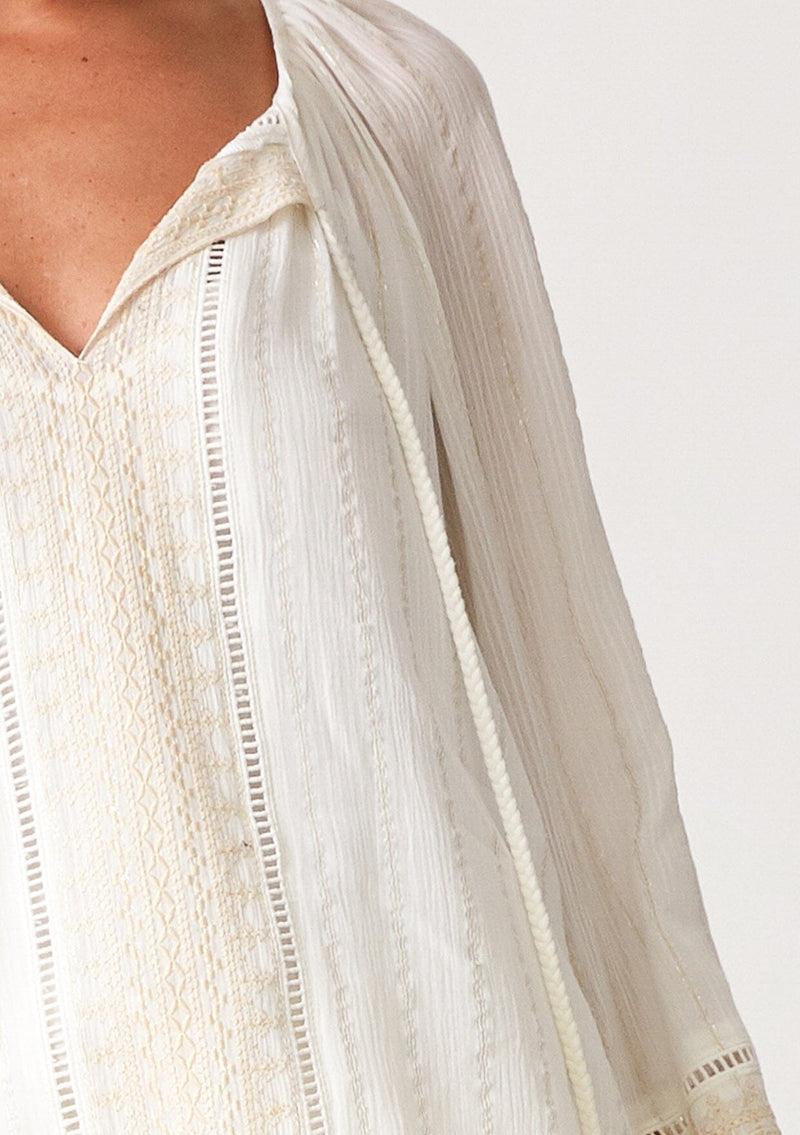 [Color: Off White] A close up front facing image of a blonde model wearing an off white bohemian fall blouse with embroidered details and sparkly metallic thread. With long raglan sleeves, a split v neckline with tassel ties, lattice trim, and a relaxed fit. 