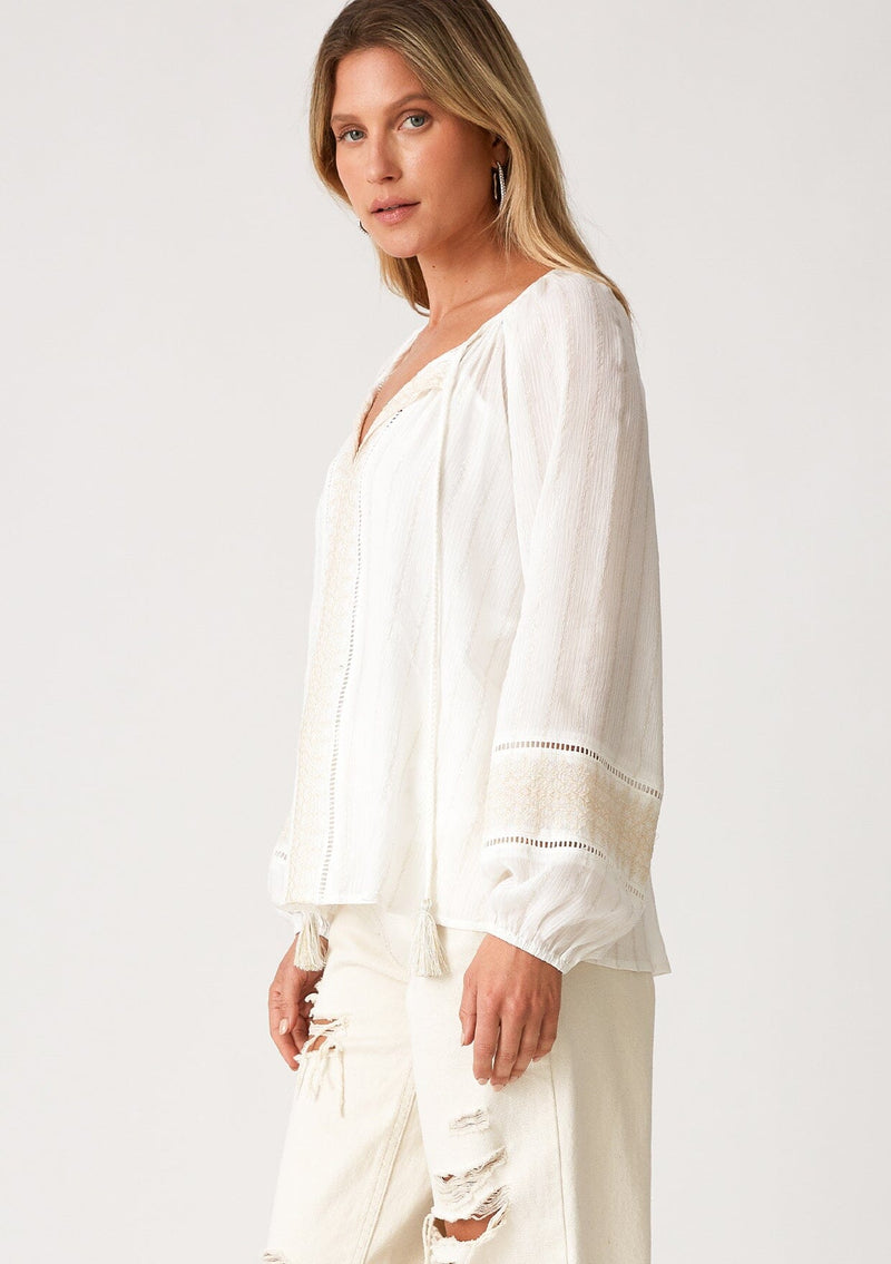 [Color: Off White] A side facing image of a blonde model wearing an off white bohemian fall blouse with embroidered details and sparkly metallic thread. With long raglan sleeves, a split v neckline with tassel ties, lattice trim, and a relaxed fit. 