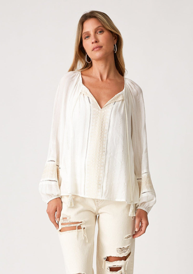 [Color: Off White] A front facing image of a blonde model wearing an off white bohemian fall blouse with embroidered details and sparkly metallic thread. With long raglan sleeves, a split v neckline with tassel ties, lattice trim, and a relaxed fit. 