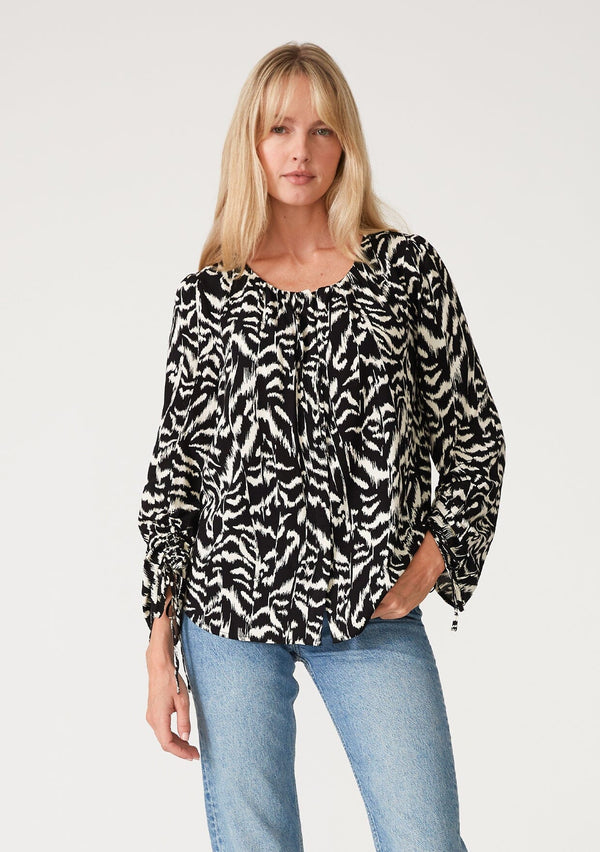 [Color: Black/Natural] A front facing image of a blonde model wearing a bohemian resort blouse in a black and off white abstract print. With long sleeves, drawstring sleeve detail, tie sleeves, a round neckline, and a concealed button front. 