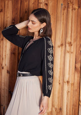 [Color: Black/Natural] A side facing image of a brunette model standing outside wearing a bohemian black blouse with embroidered detail. With voluminous long sleeves, a v neckline, and a relaxed fit.