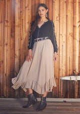[Color: Taupe] A front facing image of a brunette model standing outside wearing a taupe grey maxi length skirt. With a trendy low rise waist, a ruffled hemline, smocked elastic waist details, and a flowy silhouette.