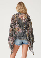 [Color: Black/Dusty Peach] A back facing image of a blonde model wearing an ultra flowy bohemian resort blouse in a black and pink mixed floral print. Crafted from chiffon and featuring long dolman sleeves, a high round neckline, side slits, and a button front. 