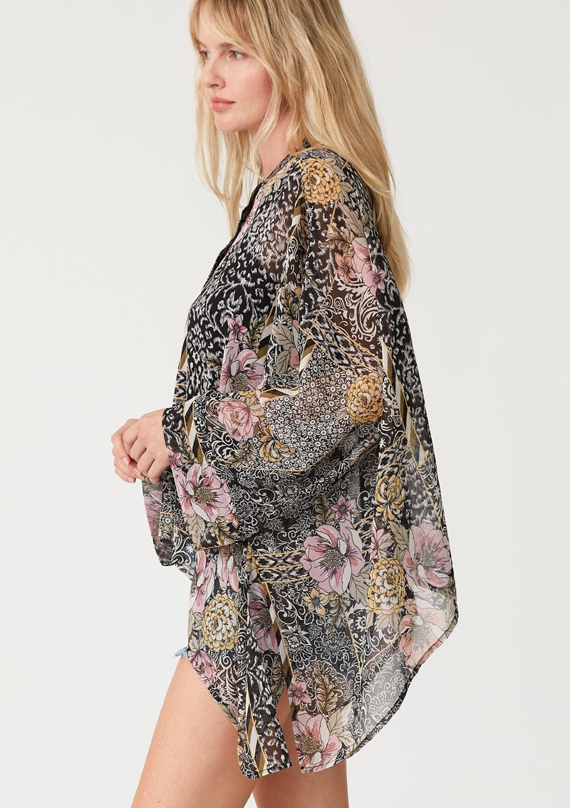 [Color: Black/Dusty Peach] A side facing image of a blonde model wearing an ultra flowy bohemian resort blouse in a black and pink mixed floral print. Crafted from chiffon and featuring long dolman sleeves, a high round neckline, side slits, and a button front. 