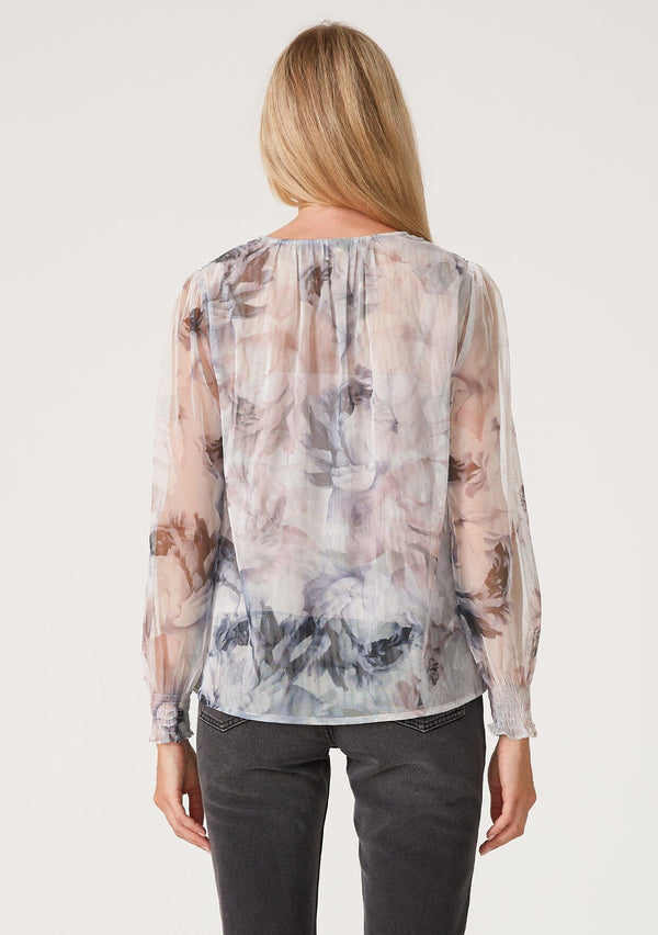 [Color: Grey/Dusty Rust] A back facing image of a blonde model wearing a bohemian resort blouse in sheer mesh tulle, designed in a grey and dusty rust floral print. With long sleeves, smocked elastic wrist cuffs, and a v neckline. 