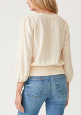 [Color: Cream] A back facing image of a blonde model wearing a cream bohemian resort top with three quarter length sleeves, a surplice v neckline with lace trim, and a smocked elastic waist. 