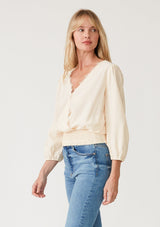 [Color: Cream] A side facing image of a blonde model wearing a cream bohemian resort top with three quarter length sleeves, a surplice v neckline with lace trim, and a smocked elastic waist. 