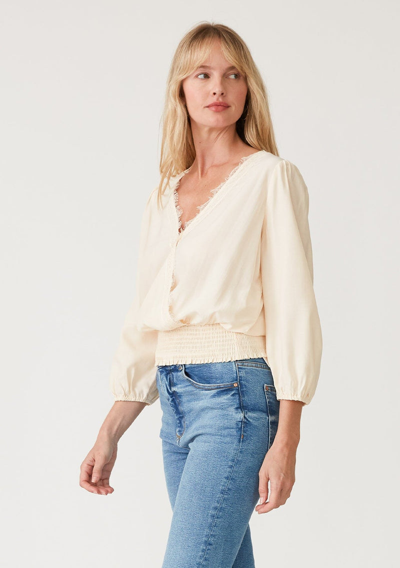 [Color: Cream] A side facing image of a blonde model wearing a cream bohemian resort top with three quarter length sleeves, a surplice v neckline with lace trim, and a smocked elastic waist. 