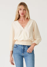 [Color: Cream] A front facing image of a blonde model wearing a cream bohemian resort top with three quarter length sleeves, a surplice v neckline with lace trim, and a smocked elastic waist. 