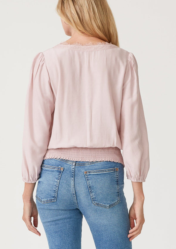 [Color: Dusty Pink] A back facing image of a blonde model wearing a dusty pink bohemian resort top with three quarter length sleeves, a surplice v neckline with lace trim, and a smocked elastic waist. 