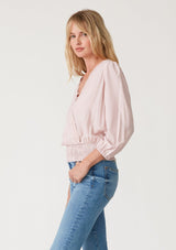[Color: Dusty Pink] A side facing image of a blonde model wearing a dusty pink bohemian resort top with three quarter length sleeves, a surplice v neckline with lace trim, and a smocked elastic waist. 