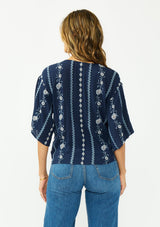 [Color: Navy/Blue] A back facing image of a brunette model wearing a blue embroidered tie front top with half length sleeves and a v neckline. 