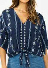 [Color: Navy/Blue] A detail image of a brunette model wearing a blue embroidered tie front top with half length sleeves and a v neckline. 