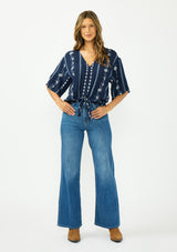 [Color: Navy/Blue] A front facing image of a brunette model wearing a blue embroidered tie front top with half length sleeves and a v neckline. Styled with a blue denim and brown boots. 