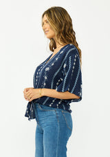 [Color: Navy/Blue] A side facing image of a brunette model wearing a blue embroidered tie front top with half length sleeves and a v neckline. 