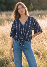 [Color: Navy/Blue] A woman stands in a sunlit field wearing a navy blue short-sleeve blouse with white vertical floral and geometric patterns, paired with light blue jeans.