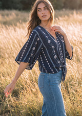 [Color: Navy/Blue] A woman wearing a navy blue bohemian-style blouse with white floral and geometric patterns, paired with light blue jeans, standing in a field of dry golden grass during golden hour.
