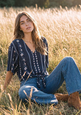 [Color: Navy/Blue] A woman sitting in a sunlit grassy field wearing a navy blue embroidered blouse with wide-leg blue jeans and brown boots.
