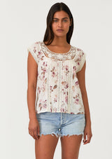 [Color: Natural/Dusty Wine] A front facing image of a brunette model wearing a bohemian resort top in a vintage inspired off white and pink floral print. With a scoop neckline, short cap sleeves, lace trim, pintuck details, and a self covered button up back. 