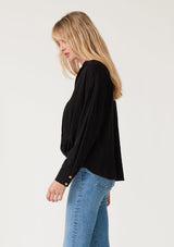 [Color: Black] A side facing image of a blonde model wearing a black linen blend relaxed button up shirt. With long sleeves, a dropped shoulder, a collared v neckline, a high low hemline, and a knot front waist detail.