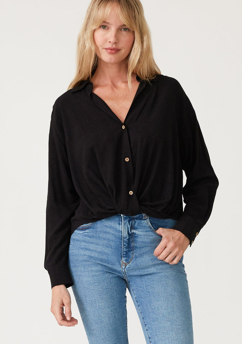 [Color: Black] A front facing image of a blonde model wearing a black linen blend relaxed button up shirt. With long sleeves, a dropped shoulder, a collared v neckline, a high low hemline, and a knot front waist detail.