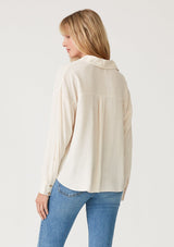 [Color: Vanilla] A back facing image of a blonde model wearing an ivory linen blend relaxed button up shirt. With long sleeves, a dropped shoulder, a collared v neckline, a high low hemline, and a knot front waist detail. 