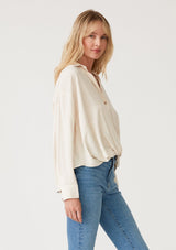 [Color: Vanilla] A side facing image of a blonde model wearing an ivory linen blend relaxed button up shirt. With long sleeves, a dropped shoulder, a collared v neckline, a high low hemline, and a knot front waist detail. 