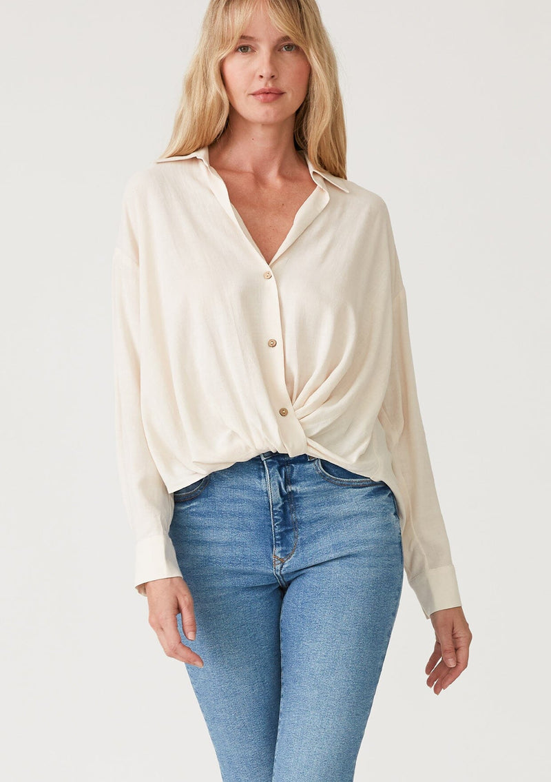 [Color: Vanilla] A front facing image of a blonde model wearing an ivory linen blend relaxed button up shirt. With long sleeves, a dropped shoulder, a collared v neckline, a high low hemline, and a knot front waist detail. 