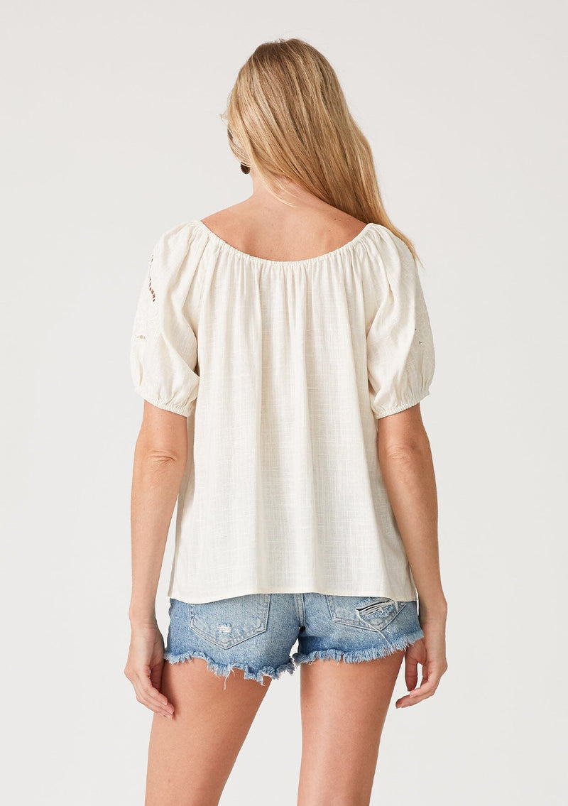 [Color: Vanilla] A back facing image of a blonde model wearing an off white bohemian resort top with embroidered details. With short puff sleeves and a wide scooped neckline with a drawstring and tassel ties. 