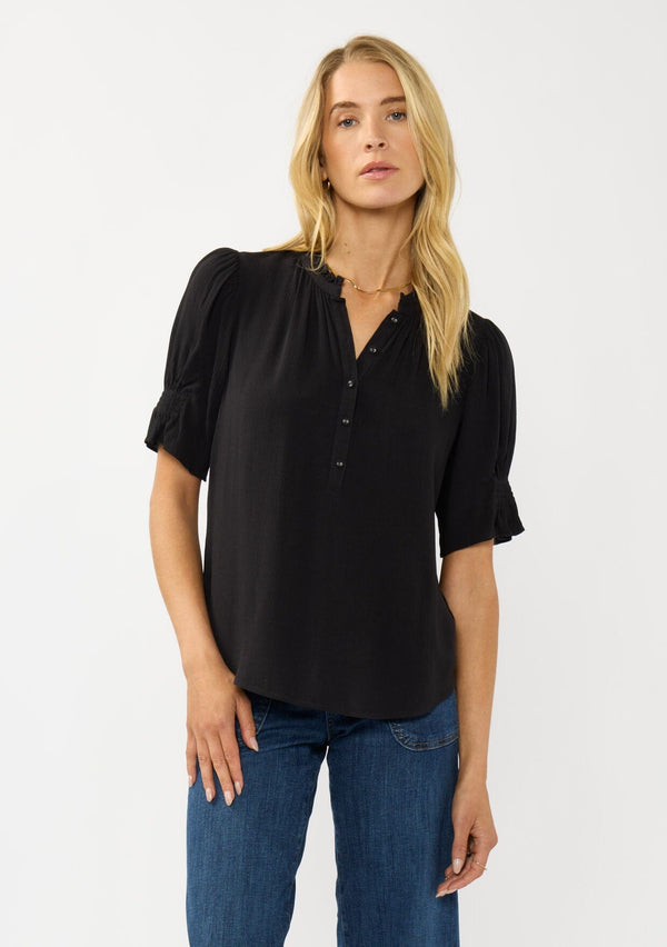 [Color: Black] A front facing image of a blonde model wearing a casual black puff sleeve top with a ruffled neckline, button front, and a gathered sleeve detail. Effortless and versatile for easy layering or wearing on its own. 