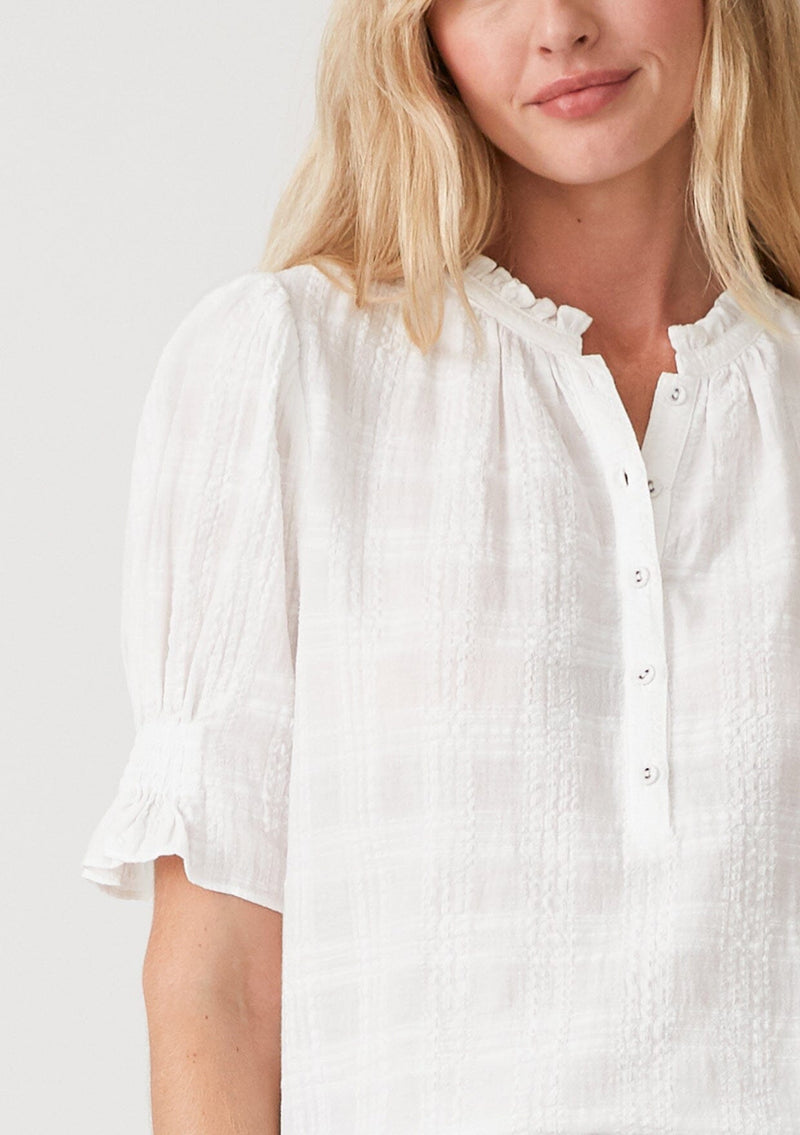 [Color: White] A close up front facing image of a blonde model wearing a white bohemian cotton blend blouse in a textured gingham. With short puff sleeves, a button front, a high ruffled neckline, and smocked elastic details at the sleeve.