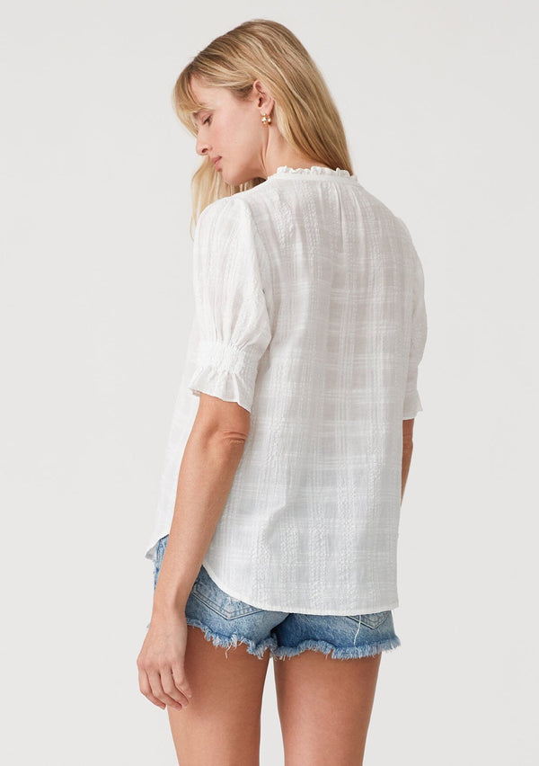 [Color: White] A back facing image of a blonde model wearing a white bohemian cotton blend blouse in a textured gingham. With short puff sleeves, a button front, a high ruffled neckline, and smocked elastic details at the sleeve. 