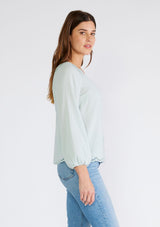 [Color: Dusty Seafoam] A side facing image of a brunette model wearing a feminine spring blouse in dusty seafoam. With scalloped embroidered lace trim, a button front, a v neckline, and long sleeves with elastic wrist cuffs. 