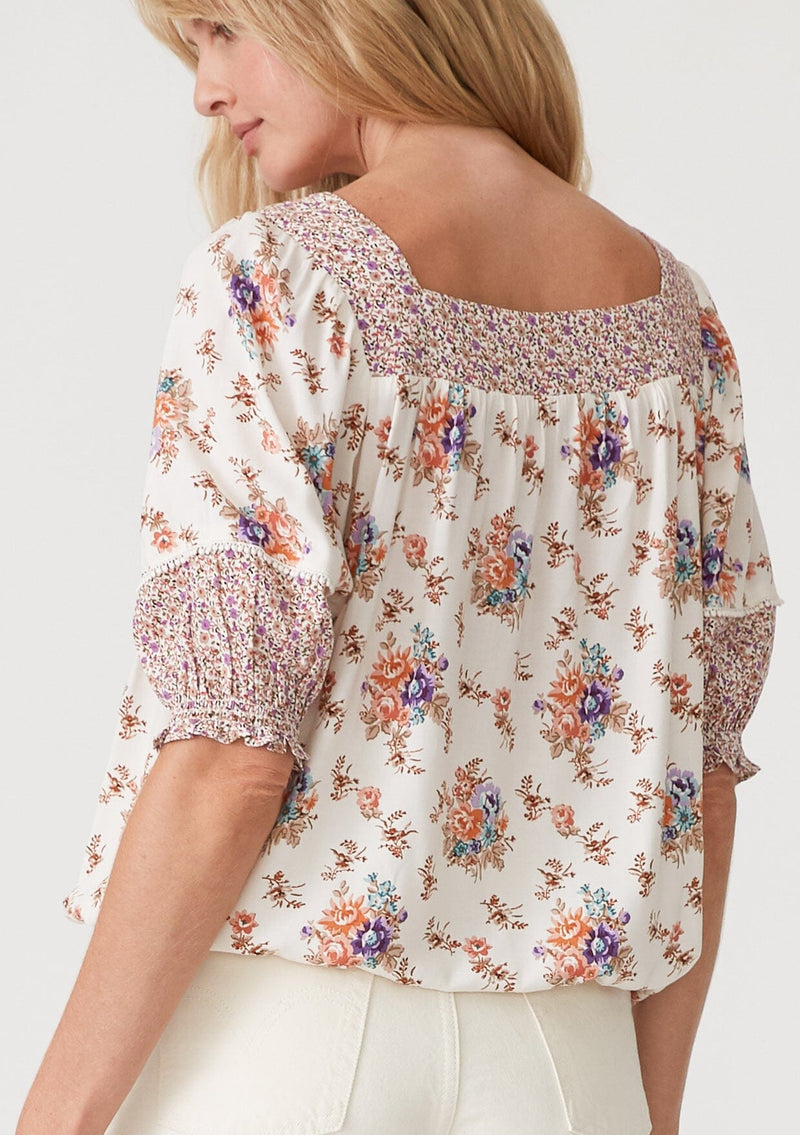[Color: Natural/Purple] A back facing image of a blonde model wearing a bohemian resort blouse in an off white and purple floral border print. With short puff sleeves, a square neckline, a button front, an adjustable drawstring waist, and a mini pom trim. 