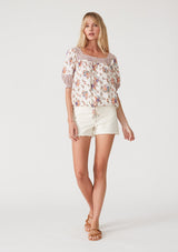 [Color: Natural/Purple] A full body front facing image of a blonde model wearing a bohemian resort blouse in an off white and purple floral border print. With short puff sleeves, a square neckline, a button front, an adjustable drawstring waist, and a mini pom trim. 