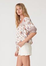 [Color: Natural/Purple] A side facing image of a blonde model wearing a bohemian resort blouse in an off white and purple floral border print. With short puff sleeves, a square neckline, a button front, an adjustable drawstring waist, and a mini pom trim. 