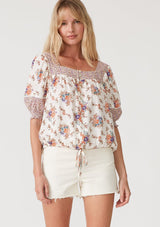 [Color: Natural/Purple] A front facing image of a blonde model wearing a bohemian resort blouse in an off white and purple floral border print. With short puff sleeves, a square neckline, a button front, an adjustable drawstring waist, and a mini pom trim. 