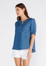 [Color: Vintage Wash] A front facing image of a brunette model wearing a denim blue spring blouse crafted from Tencel. With short puff sleeves, a round neckline, a self covered button front, a front patch pocket, and a relaxed fit. 