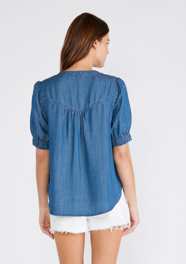 [Color: Vintage Wash] A back facing image of a brunette model wearing a denim blue spring blouse crafted from Tencel. With short puff sleeves, a round neckline, a self covered button front, a front patch pocket, and a relaxed fit. 