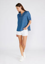 [Color: Vintage Wash] A full body front facing image of a brunette model wearing a denim blue spring blouse crafted from Tencel. With short puff sleeves, a round neckline, a self covered button front, a front patch pocket, and a relaxed fit. 