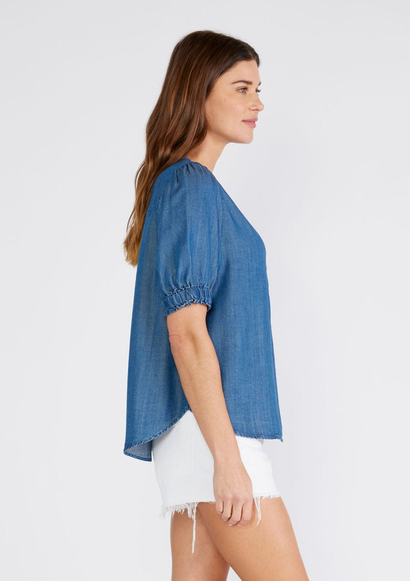 [Color: Vintage Wash] A side facing image of a brunette model wearing a denim blue spring blouse crafted from Tencel. With short puff sleeves, a round neckline, a self covered button front, a front patch pocket, and a relaxed fit. 