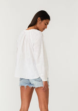 [Color: Natural] A back facing image of a brunette model wearing a white cotton bohemian blouse with long raglan sleeves, a split v neckline with ties and ruffled trim, embroidered detail, a curved hemline, and sheer lattice trim details. 
