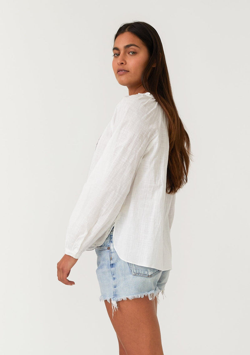 [Color: Natural] A half body side facing image of a brunette model wearing a white cotton bohemian blouse with long raglan sleeves, a split v neckline with ties and ruffled trim, embroidered detail, a curved hemline, and sheer lattice trim details. 