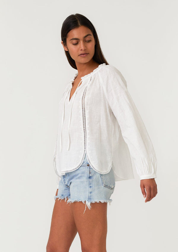 [Color: Natural] A side facing image of a brunette model wearing a white cotton bohemian blouse with long raglan sleeves, a split v neckline with ties and ruffled trim, embroidered detail, a curved hemline, and sheer lattice trim details. 