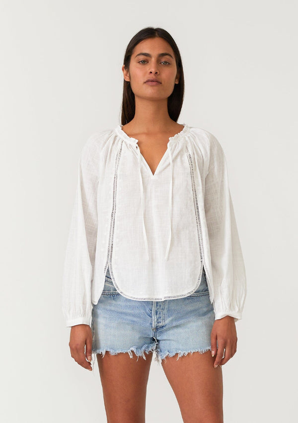 [Color: Natural] A front facing image of a brunette model wearing a white cotton bohemian blouse with long raglan sleeves, a split v neckline with ties and ruffled trim, embroidered detail, a curved hemline, and sheer lattice trim details. 
